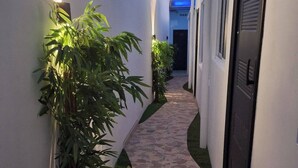 Un stretto sentiero in pietra costeggiato da pareti bianche e piante verdi, che conduce a un edificio con una porta scura e una finestra illuminata di blu.