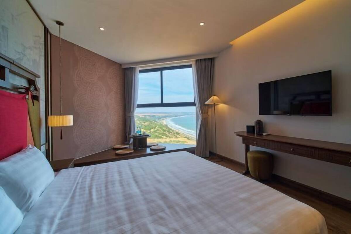 A hotel room with a large bed, a desk, a chair, a TV, and a view of the beach.