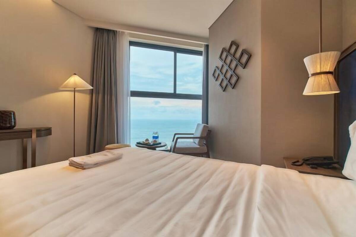 Une chambre d’hôtel avec un grand lit, un bureau avec une lampe, une chaise, une petite table avec une boisson, et une vue sur l’océan.
