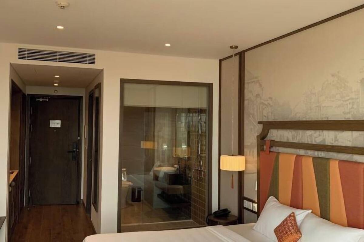 Une chambre d’hôtel avec un lit, une table de chevet, une lampe et une vue sur la salle de bain, accessible par une porte vitrée.