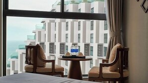 Une chambre d’hôtel avec un lit, deux chaises, une petite table ronde sur laquelle se trouvent une bouteille et des verres, et une vue sur des immeubles modernes et la mer.