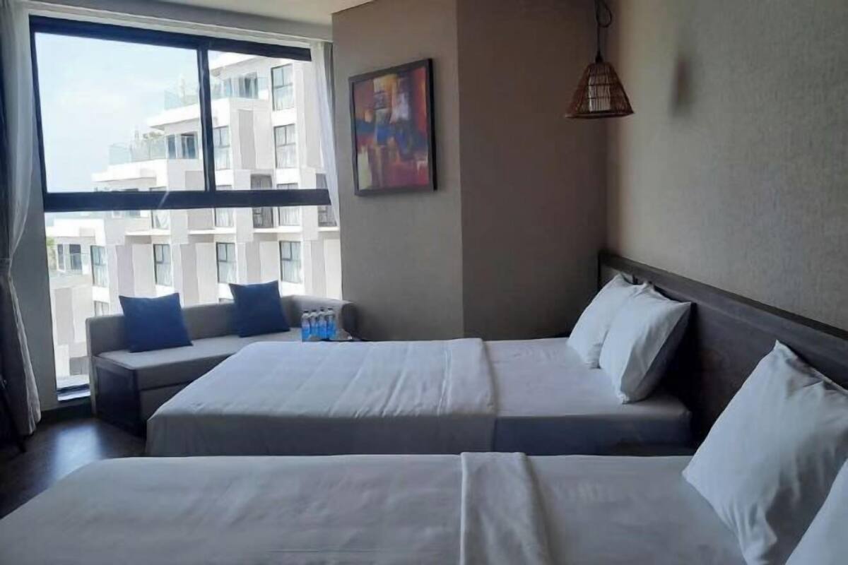 Une chambre d’hôtel avec deux lits, une fenêtre donnant sur des immeubles, un tableau accroché au mur et un luminaire suspendu.