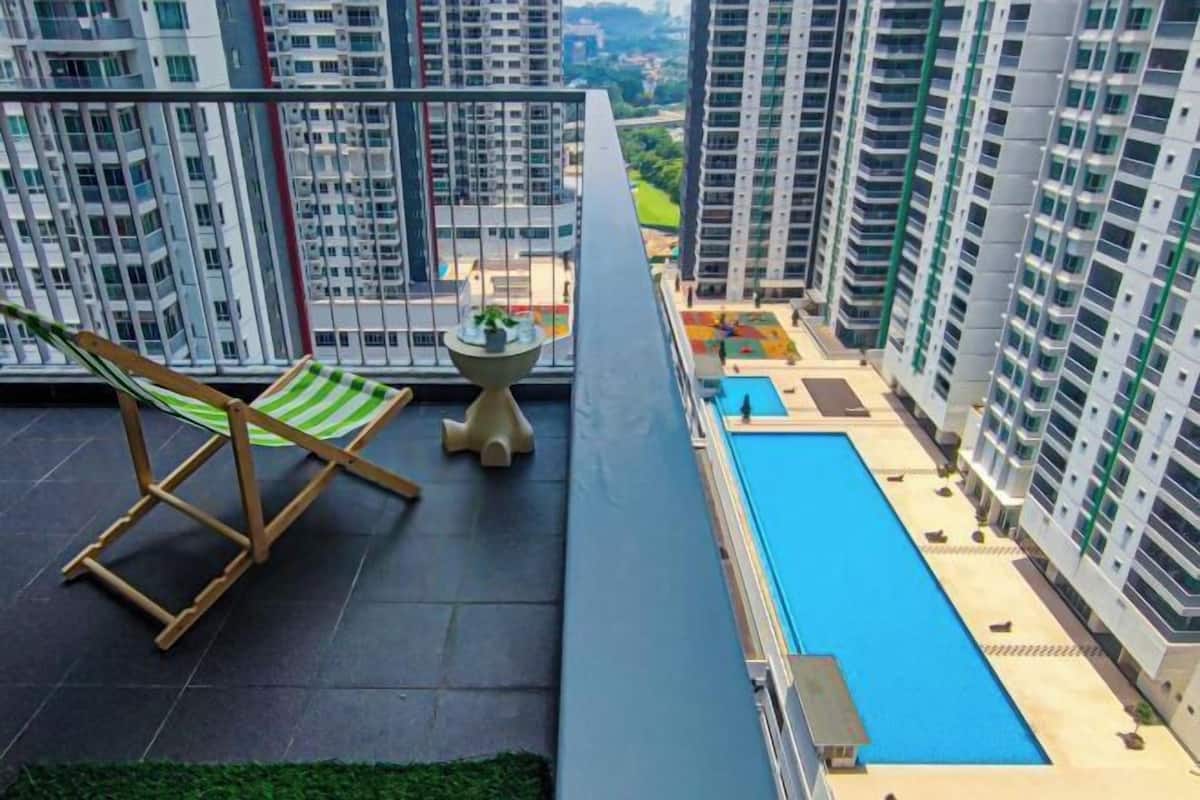 A balcony with a green-striped chair, a small table, and a pool area with buildings in the background.