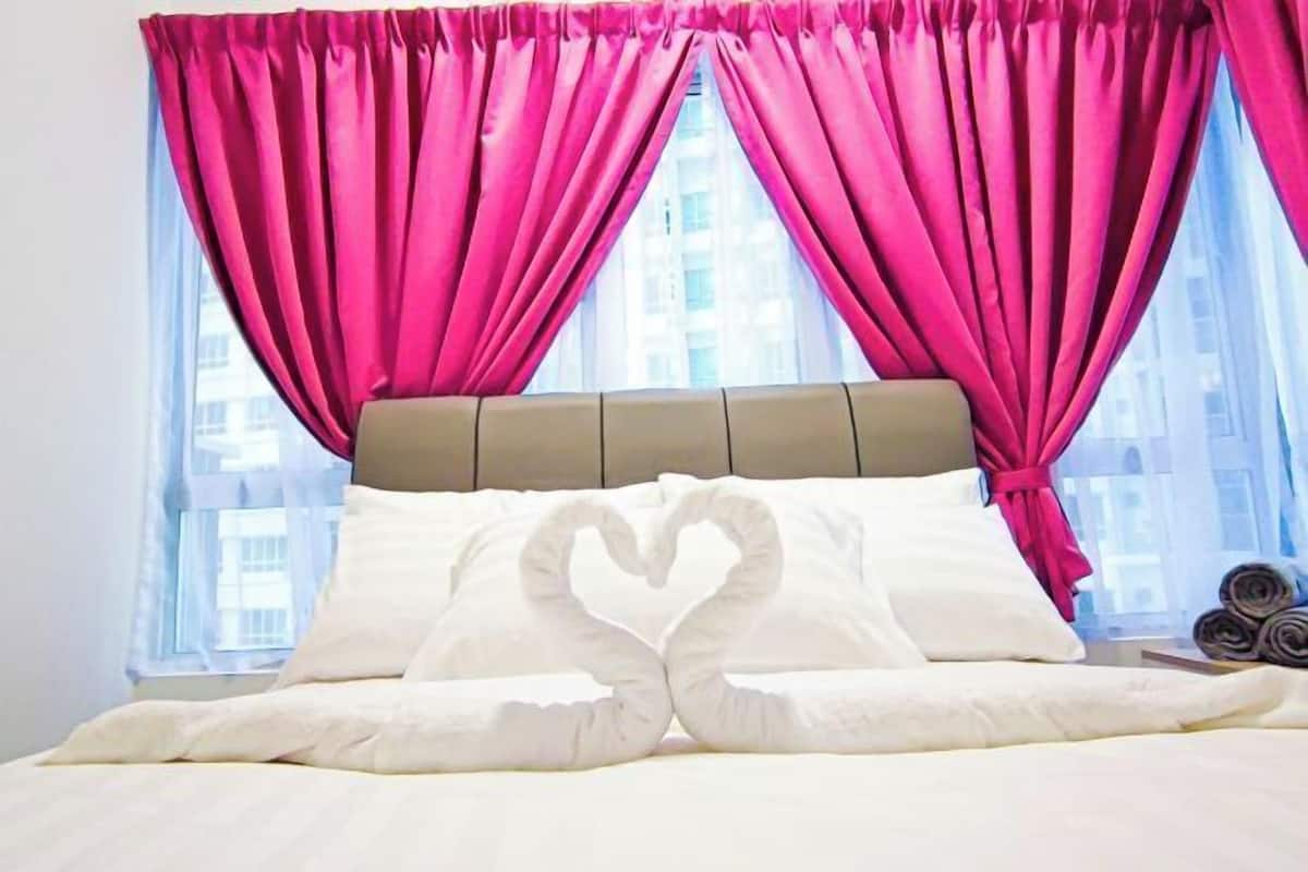 A hotel room with a bed, pink curtains, and a heart-shaped pillow arrangement.