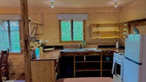 A wooden cabin interior with a kitchen area, a dining table, and a window with a view of trees. - Maccy's Cottages (Macclesfield)