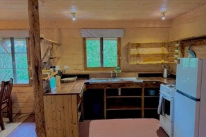 Innenansicht einer Holzhütte mit Küchenbereich, Esstisch und Fenster mit Blick auf Bäume.