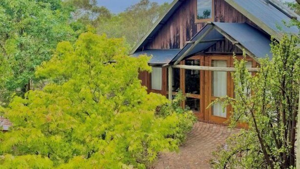 A rustic wooden house with a sloped roof, surrounded by lush greenery and a paved pathway leading to the entrance. - Maccy's Cottages (Macclesfield)