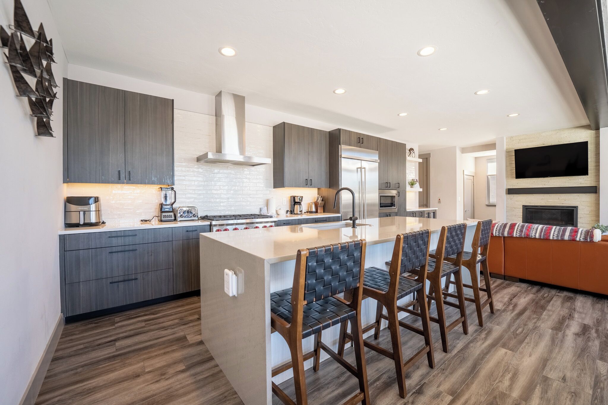 A modern kitchen with grey cabinets, a central island with bar stools, a stainless steel refrigerator, and a built-in oven.