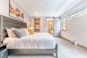 A modern bedroom with a large bed, a walk-in closet, a glass shower, and a view of the outdoors.