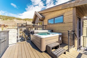 A wooden cabin with a hot tub on a deck, a stone wall, and a clear sky.