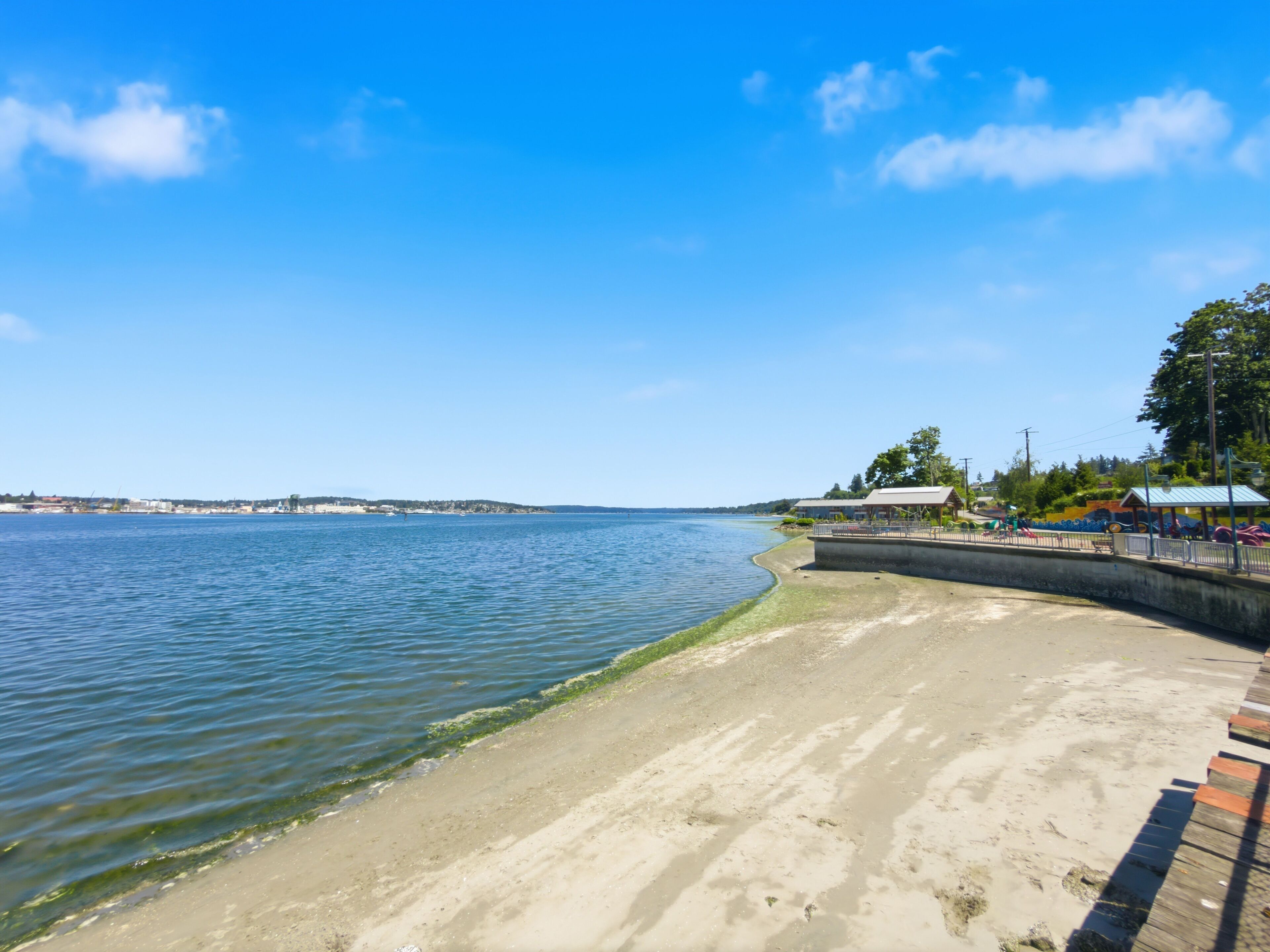 A waterfront promenade with a sandy beach, a concrete walkway, and a distant view of a cityscape.