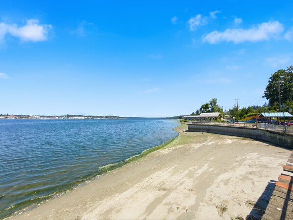 A waterfront promenade with a sandy beach, a concrete walkway, and a distant view of a cityscape.