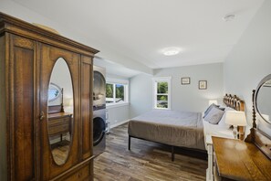 Ein Schlafzimmer mit großem Holzkoffer, einem Bett mit Kopfteil, einem Schminktisch mit Spiegel und einem Fenster mit Blick auf Grünflächen.