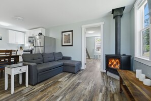 A modern living room with a grey sectional sofa, a white dining table, a black wood-burning stove, and a kitchen area with white cabinets and appliances.