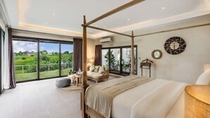 Une chambre dotée d’un grand lit, d’un cadre de lit en bois à colonnes, d’un coin salon avec un canapé et des fauteuils, et offrant une vue sur un paysage verdoyant grâce à de grandes fenêtres.