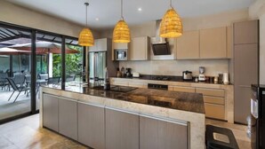 Une cuisine moderne dotée d’un îlot central, de luminaires suspendus et offrant une vue sur l’espace repas extérieur.