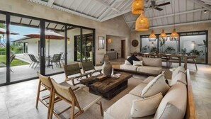 Un vaste espace de vie comprenant un grand canapé, des chaises en bois et une table basse. On y trouve également des luminaires suspendus et un ventilateur de plafond.