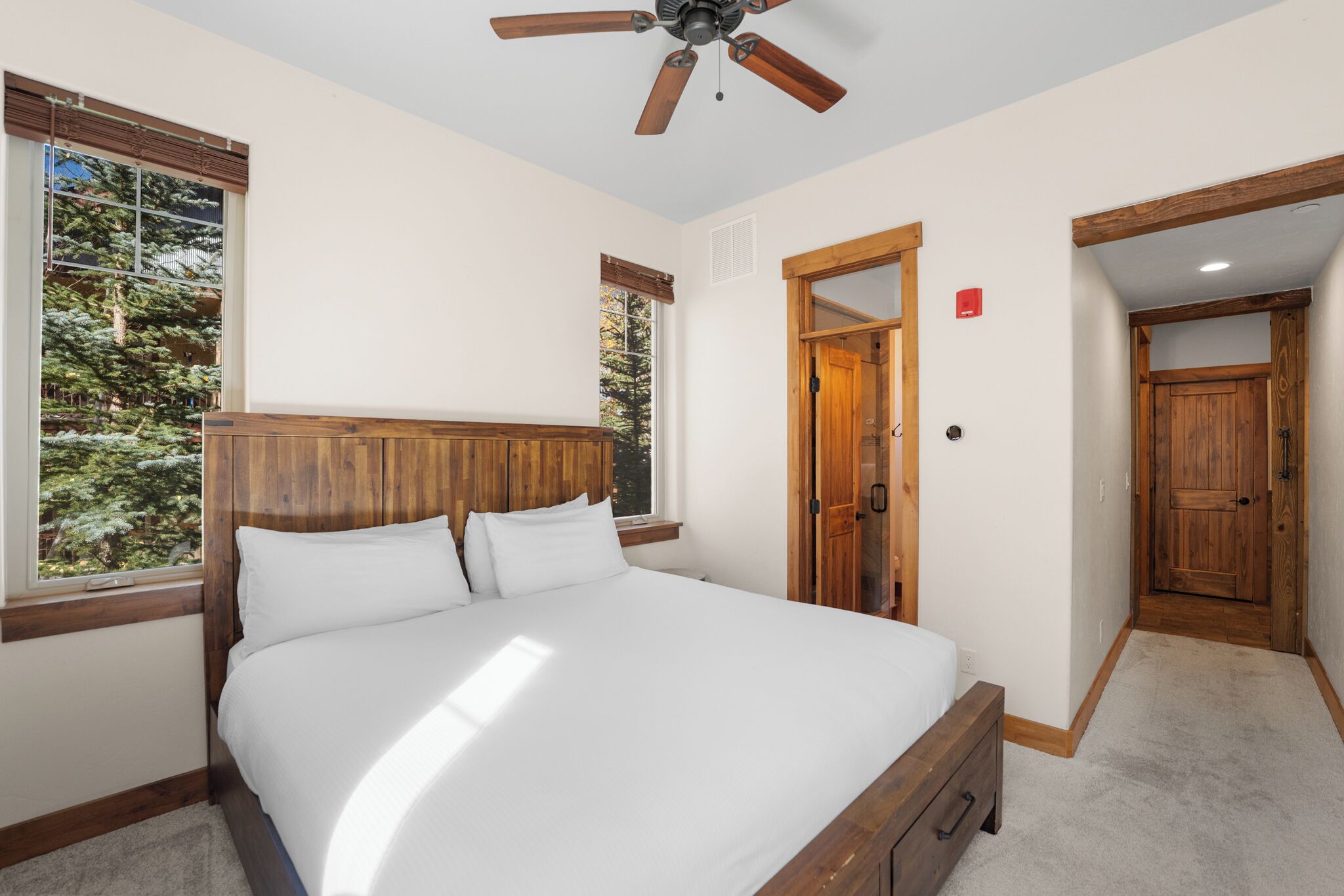 Un dormitorio con una cama grande, cabecera de madera y ventilador de techo. Hay ventanas con vistas a árboles, y una puerta de madera que da a otra habitación.