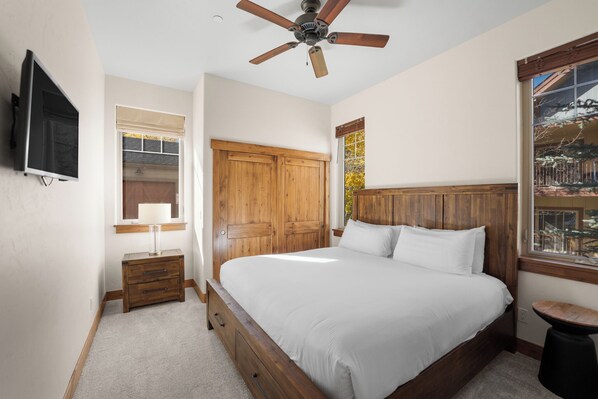 A bedroom with a large bed, wooden wardrobe, a ceiling fan, a TV mounted on the wall, and a window with a view of the outside.