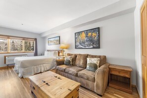 Una habitación acogedora con una cama, un sofá, una mesita de madera y una mesita de noche. Hay dos ventanas con cortinas, una lámpara y cuadros enmarcados en la pared.