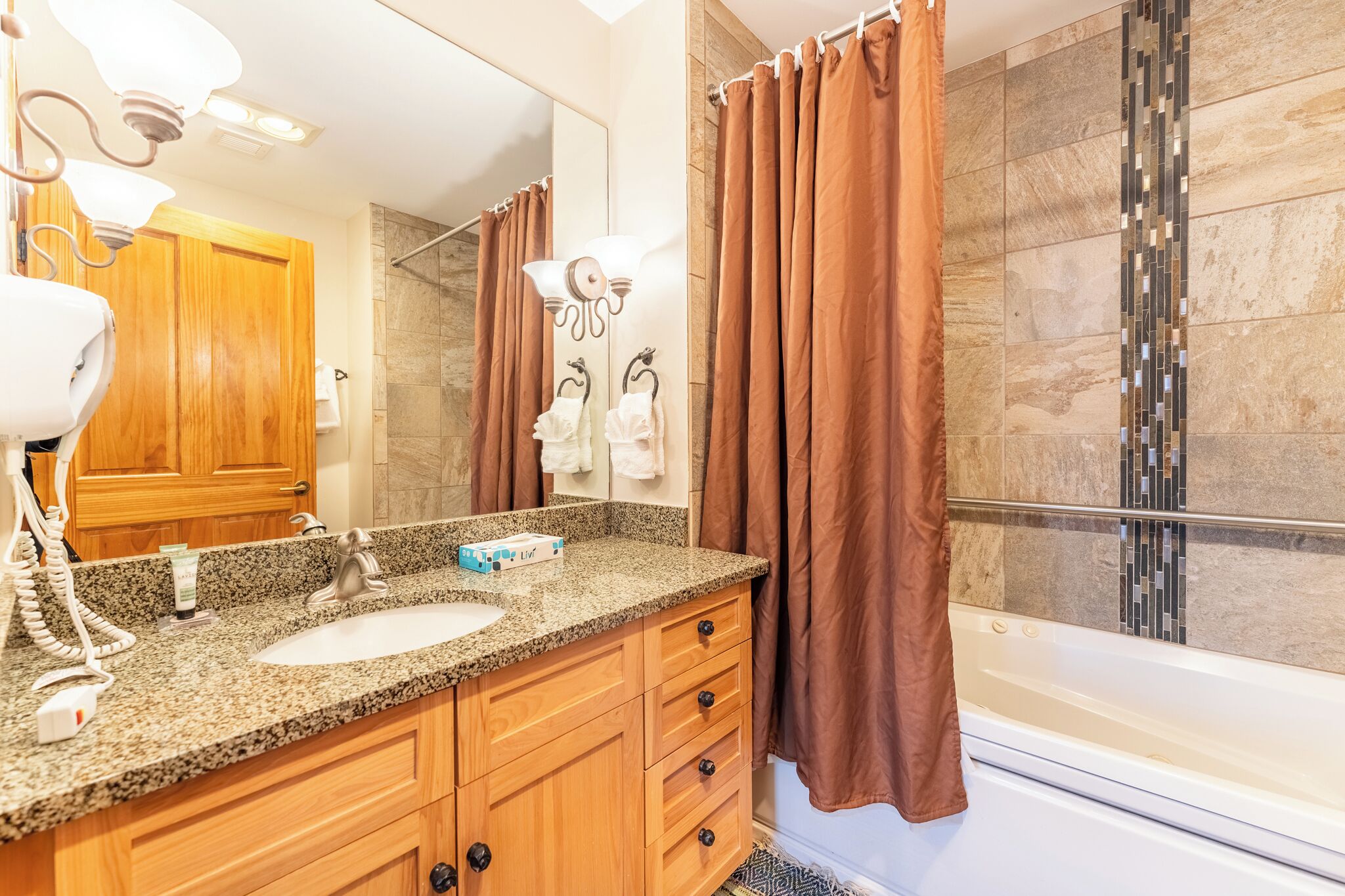 A bathroom with a granite countertop, wooden cabinets, a white sink, a mirror, a shower with a glass door, and brown curtains.