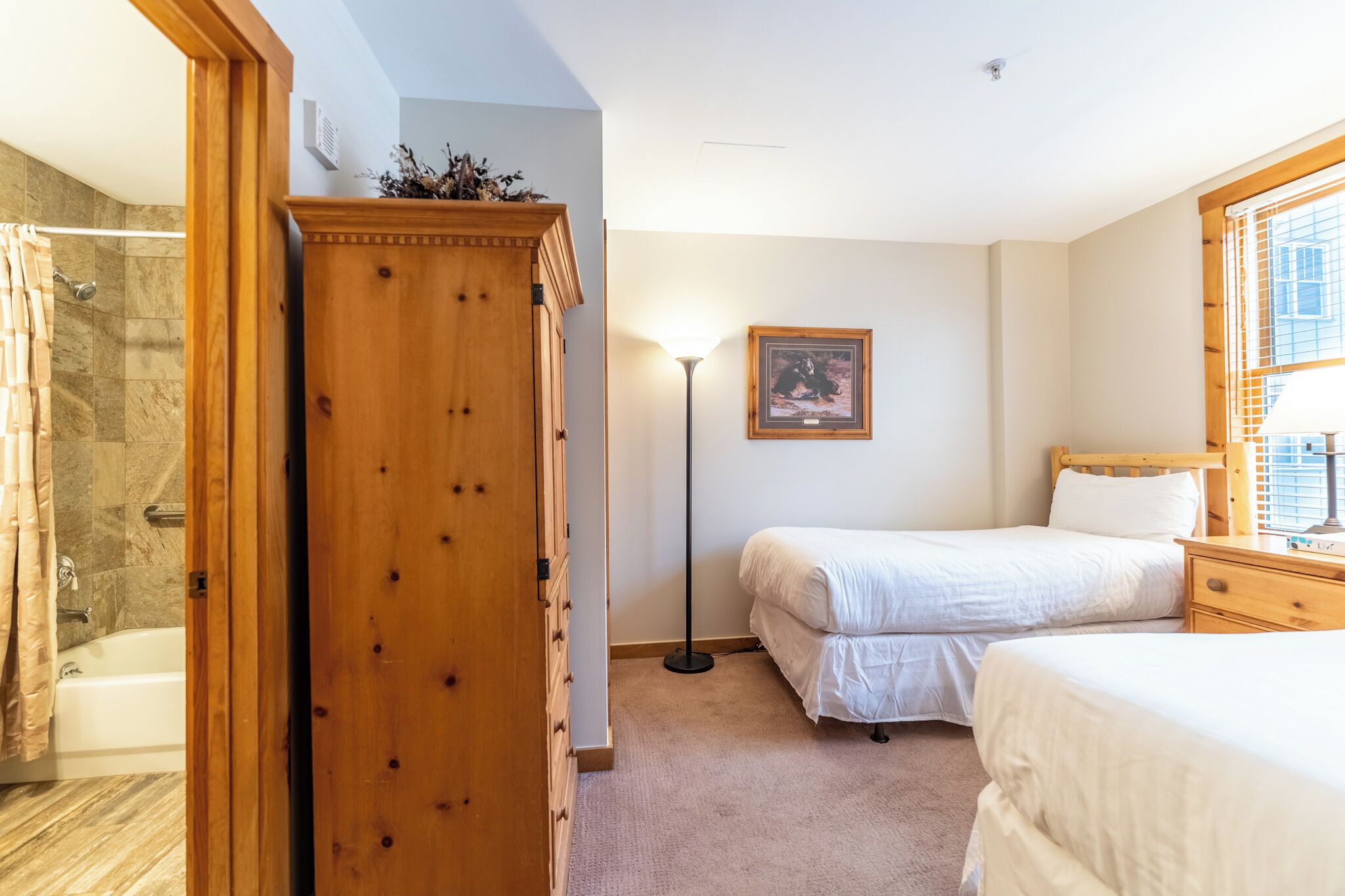 A bedroom with two beds, a wooden wardrobe, a floor lamp, and a framed picture on the wall.