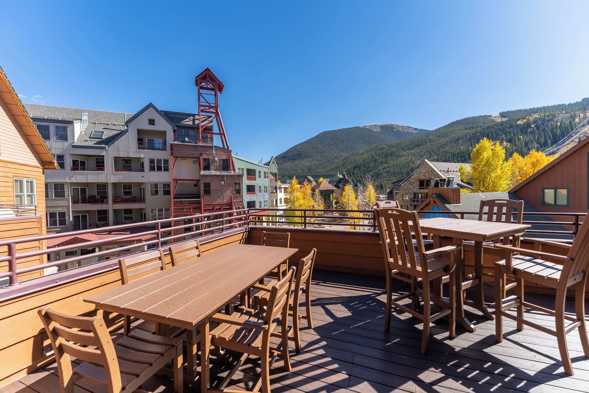 Una terraza de madera con mesas y sillas, con vistas a un paisaje montañoso y edificios al fondo.