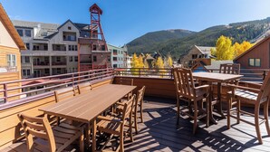 Una terraza de madera con mesas y sillas, con vistas a un paisaje montañoso y edificios al fondo.