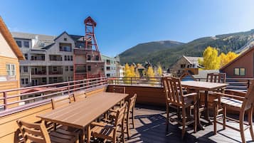 Una terraza de madera con mesas y sillas, con vistas a un paisaje montañoso y edificios al fondo.