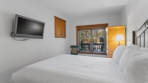 Quarto com cama, televisão fixada na parede, armário de madeira e janela com vista para árvores.
