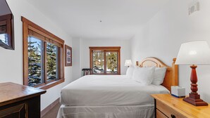 Quarto com uma cama grande, cabeceira de madeira, luminárias de cabeceira, mesa de cabeceira de madeira e uma janela com vista para árvores.