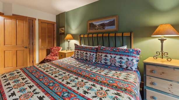Un dormitorio con una cama, dos mesitas de noche, una lámpara, una silla y un armario de madera.