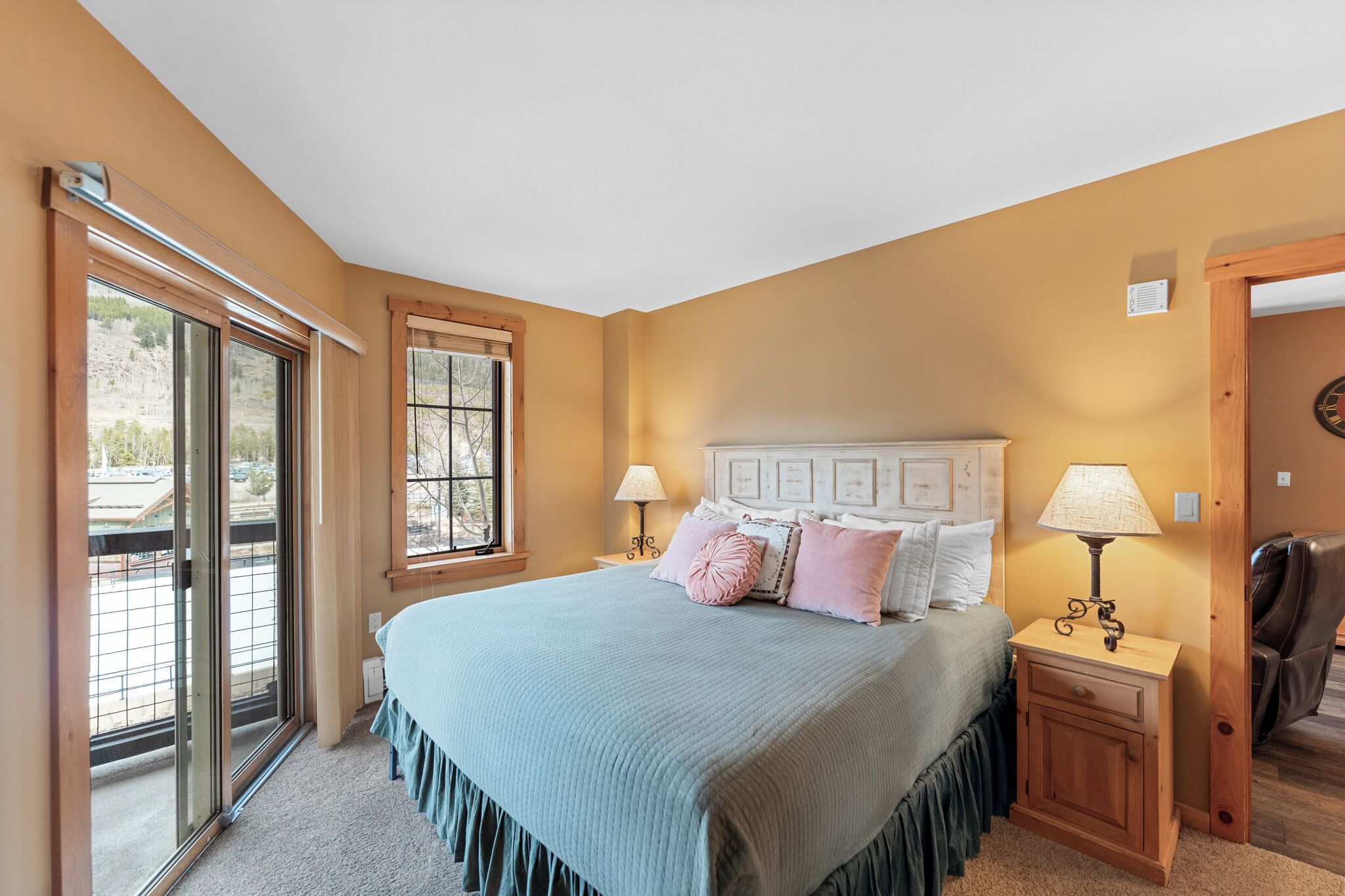 A bedroom with a large bed, two bedside tables with lamps, a wooden headboard, and a sliding glass door leading to an outdoor area.