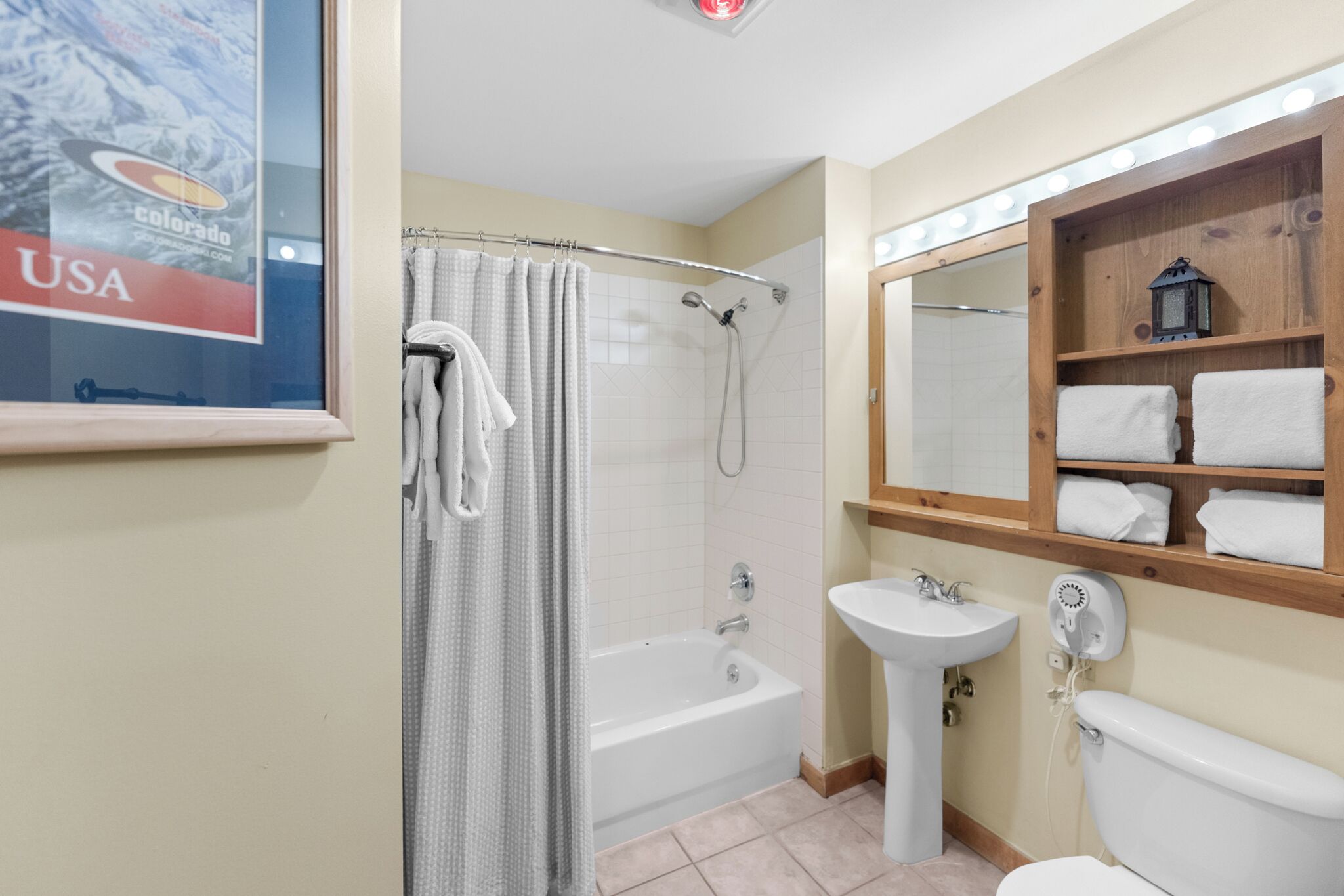 Ein Badezimmer mit Badewanne, Waschbecken und Toilette. Ein Duschvorhang, ein Handtuchhalter und ein Holzregal mit Handtüchern sind vorhanden.