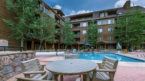 A pool area with a wooden table and chairs, surrounded by a stone wall and trees. - Dakota Lodge #8538 (Keystone)