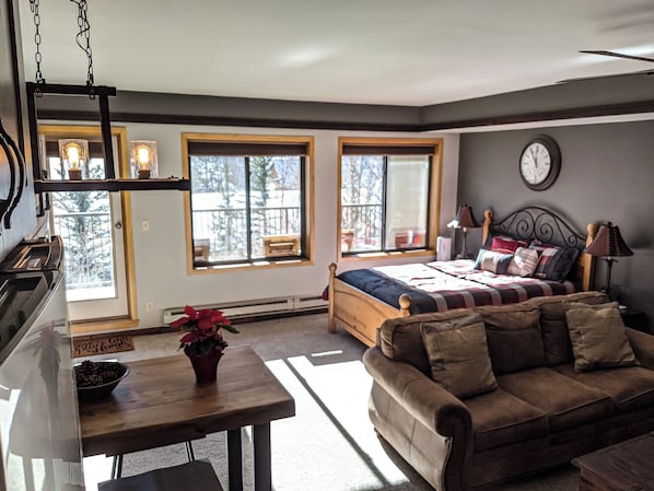 A cozy bedroom with a bed, a wooden nightstand, a bedside table with a lamp, and a view of the outdoors through large windows. - S2789-0 (Keystone)
