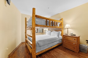 A bunk bed with a wooden frame, a bedside table with a lamp, and a trash bin.