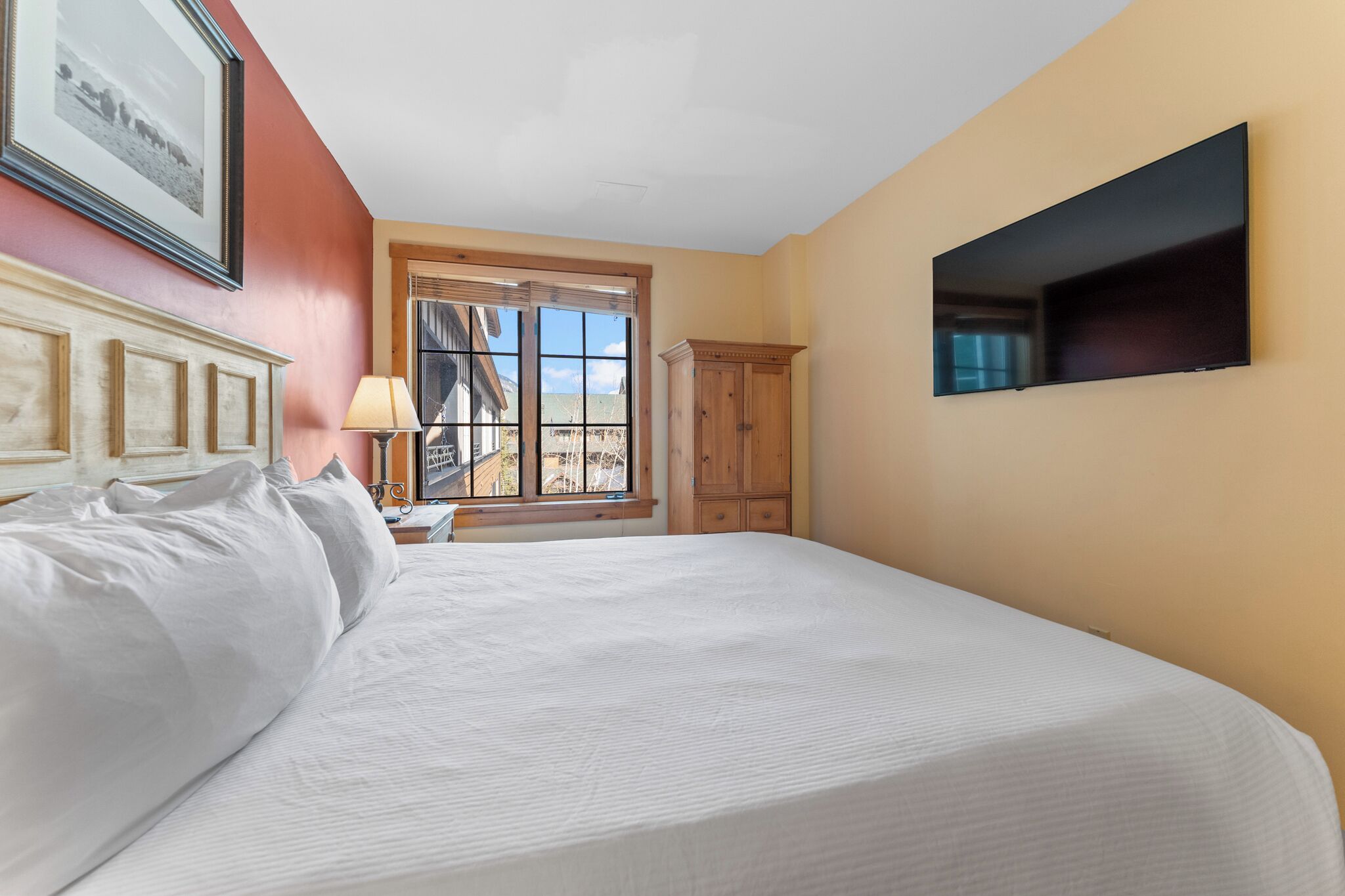 A bedroom with a large bed, a wooden wardrobe, a television mounted on the wall, and a window with a view of buildings.