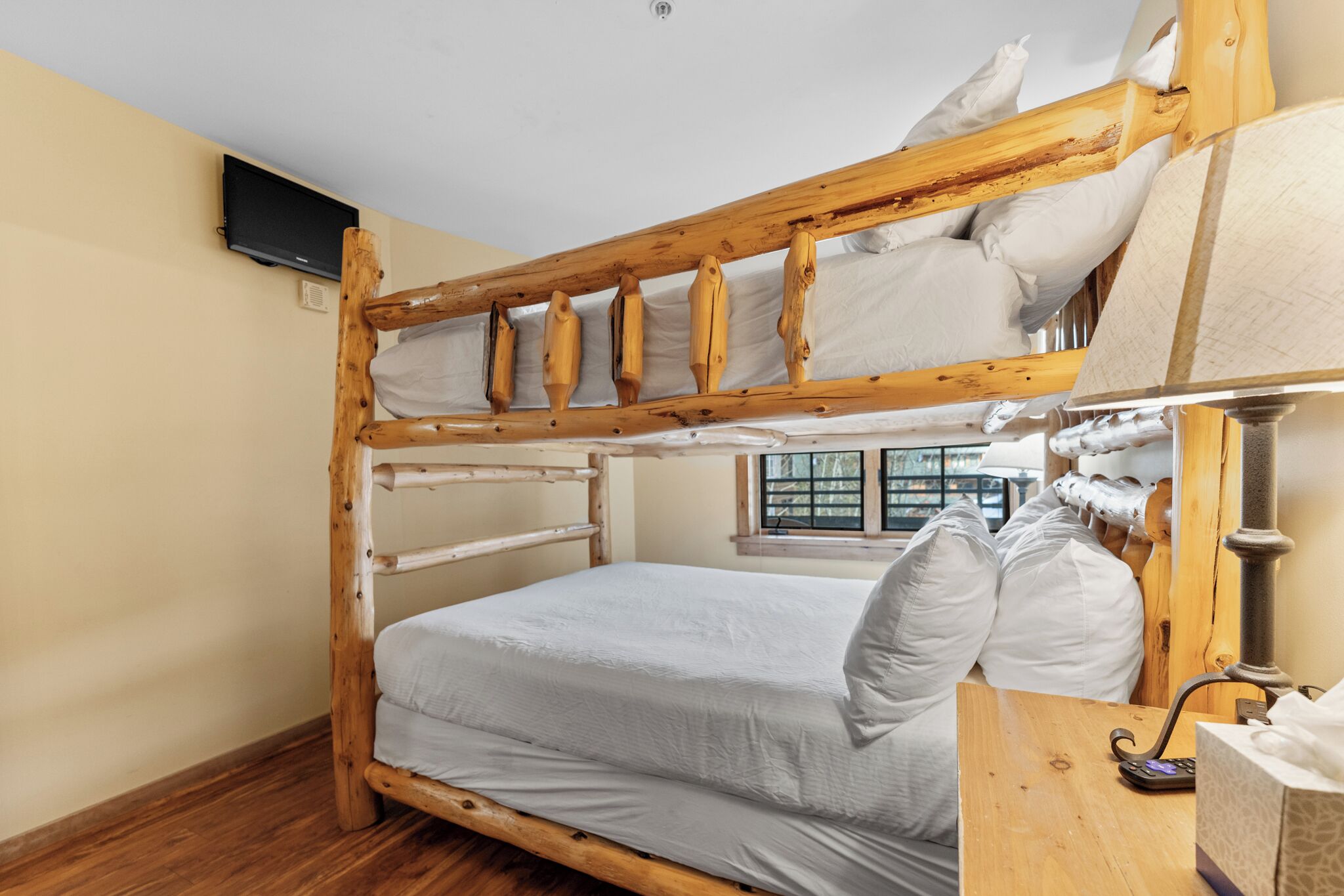Ein Etagenbett mit Holzrahmen, ein Nachttisch mit Lampe und ein Fenster mit Jalousien.