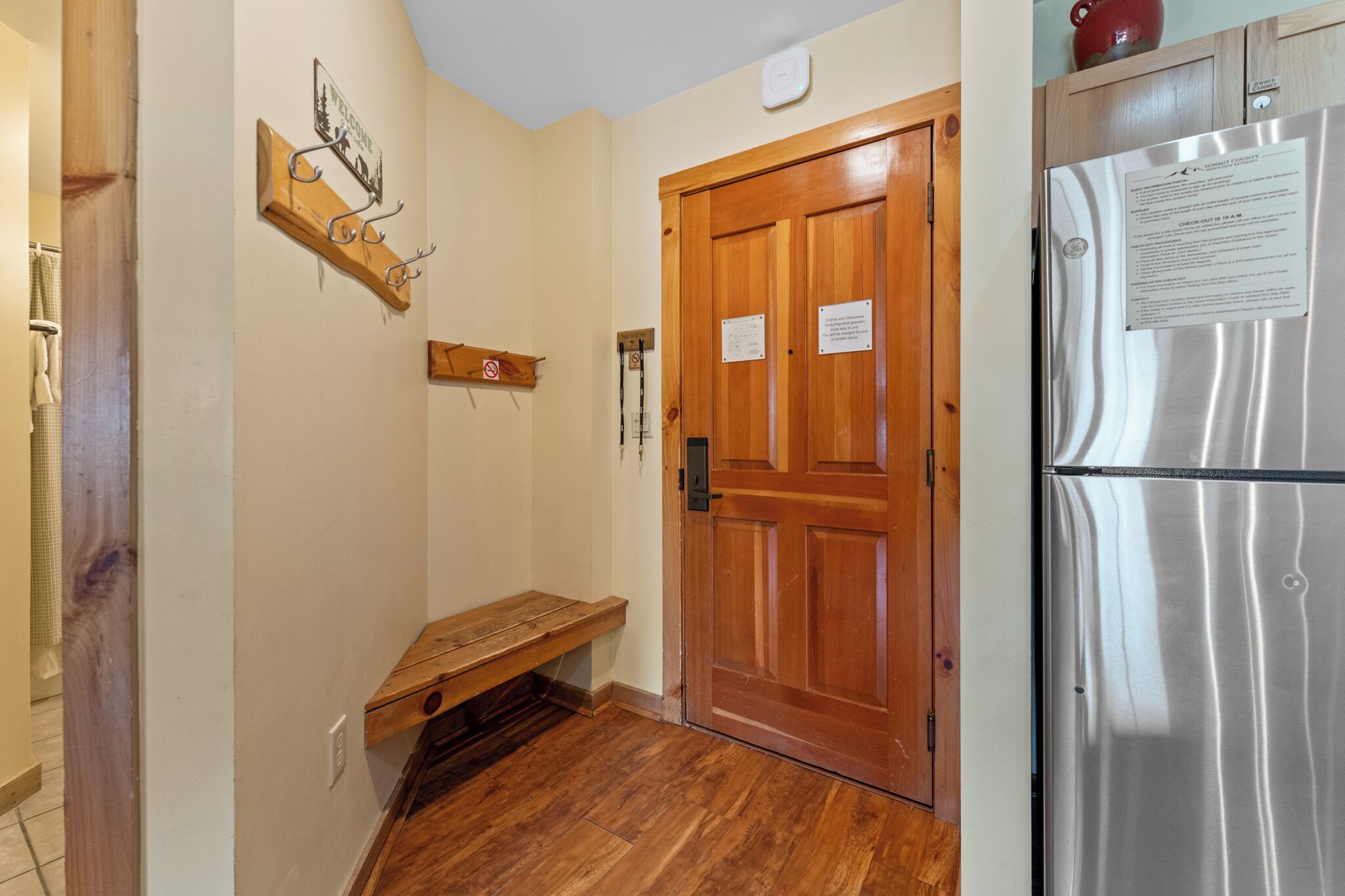 A hallway with a wooden bench, a wooden door with a sign, a stainless steel refrigerator, and a wall-mounted clock.