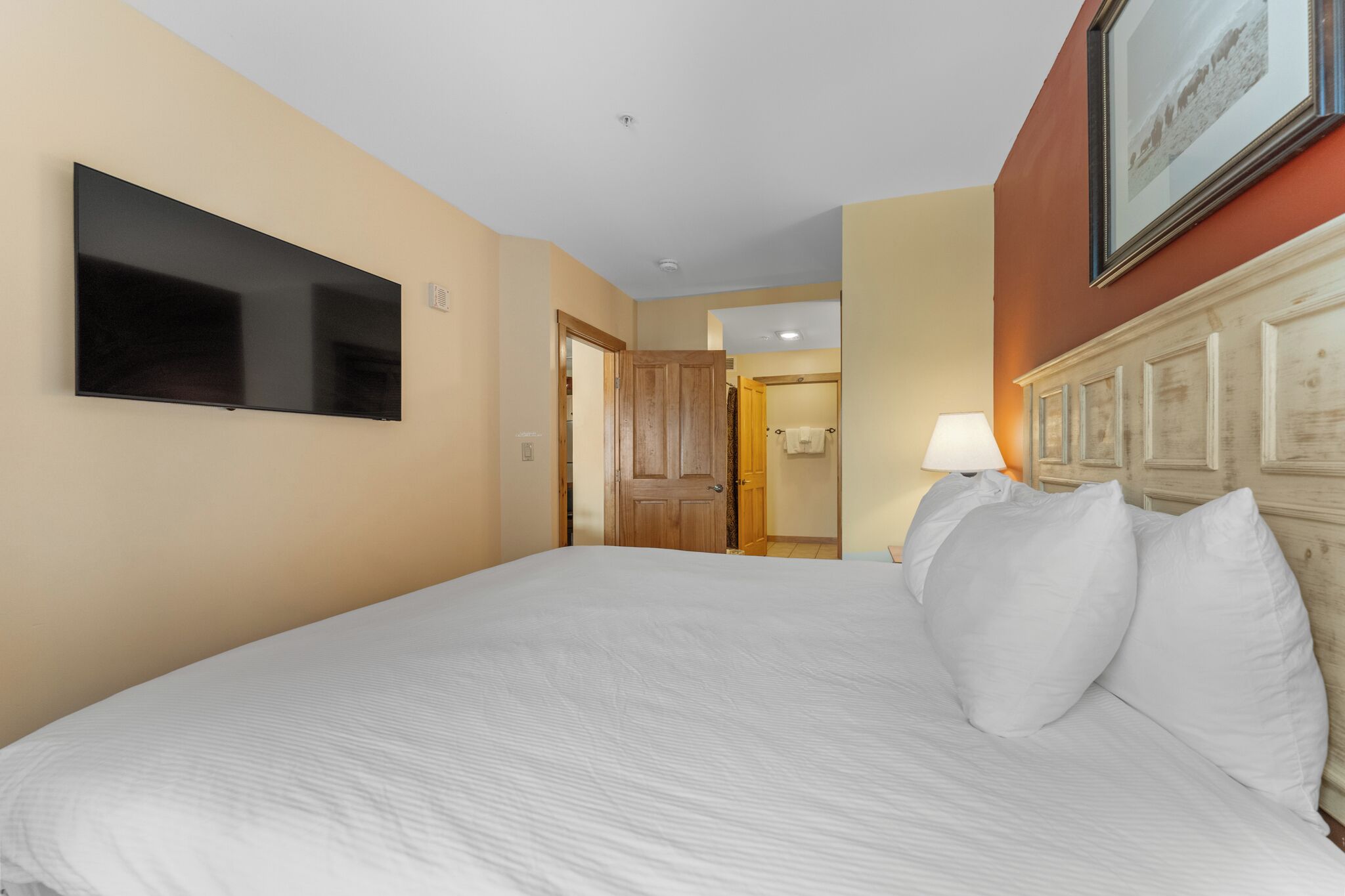 A hotel room with a large bed, a flat-screen TV mounted on the wall, a wooden door, and a framed picture above the bed.
