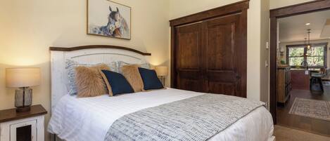 A bedroom with a large bed, a wooden wardrobe, a nightstand with a lamp, and a framed picture on the wall.