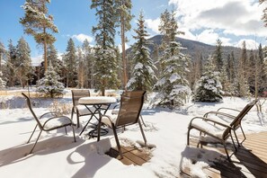 An outdoor seating area with metal chairs and a table, surrounded by snow-covered trees and a mountain in the background.