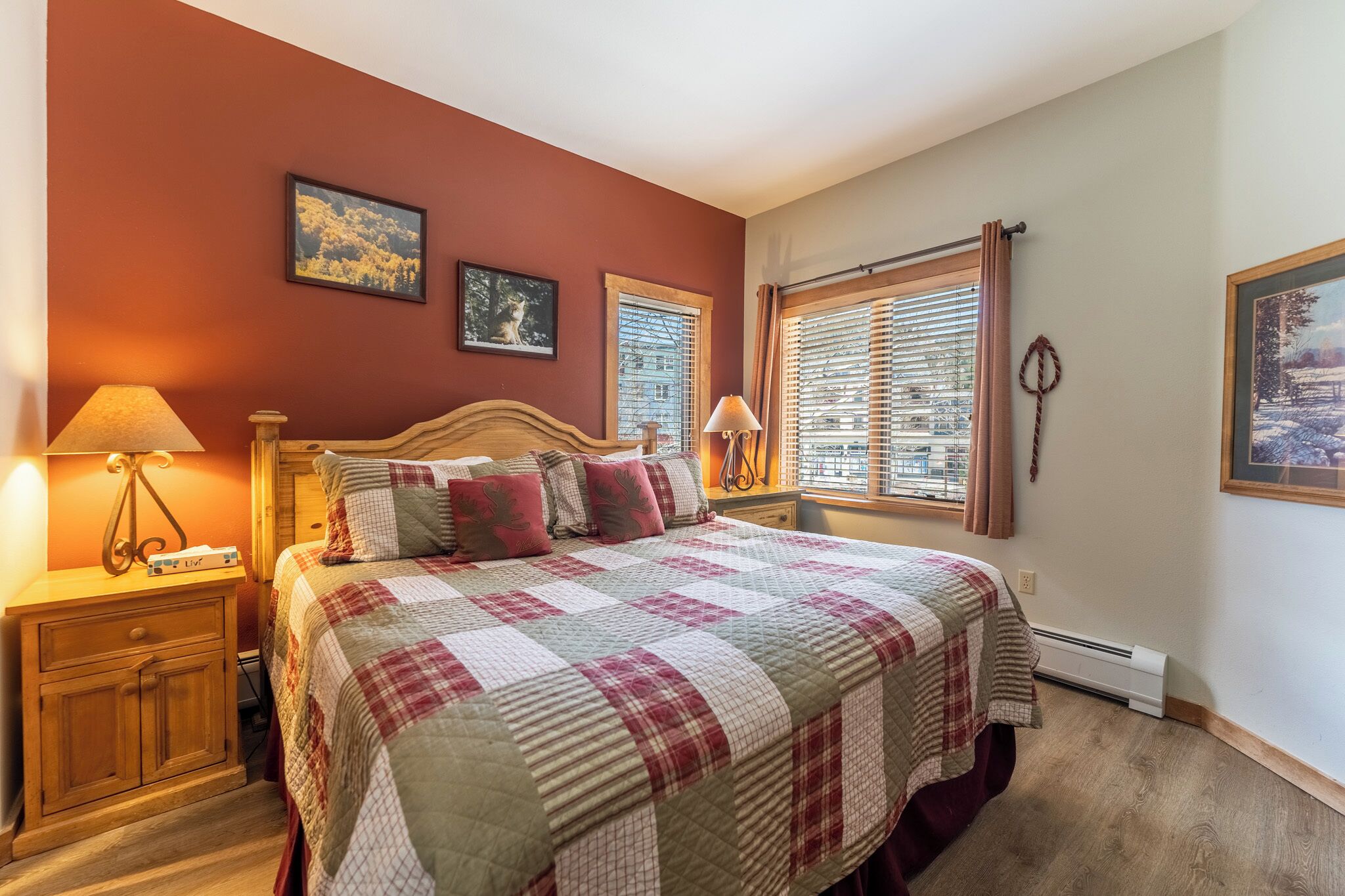 A bedroom with a bed, two bedside tables, a nightstand, a window with blinds, and two wall-mounted pictures.