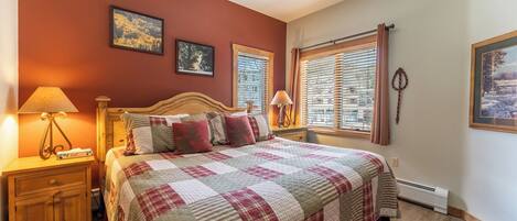 A bedroom with a bed, two bedside tables, a nightstand, a window with blinds, and two wall-mounted pictures.