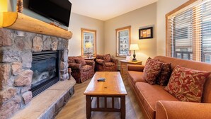 A cozy living room with a stone fireplace, a flat-screen TV mounted above it, a wooden coffee table, and a red sofa with patterned cushions.