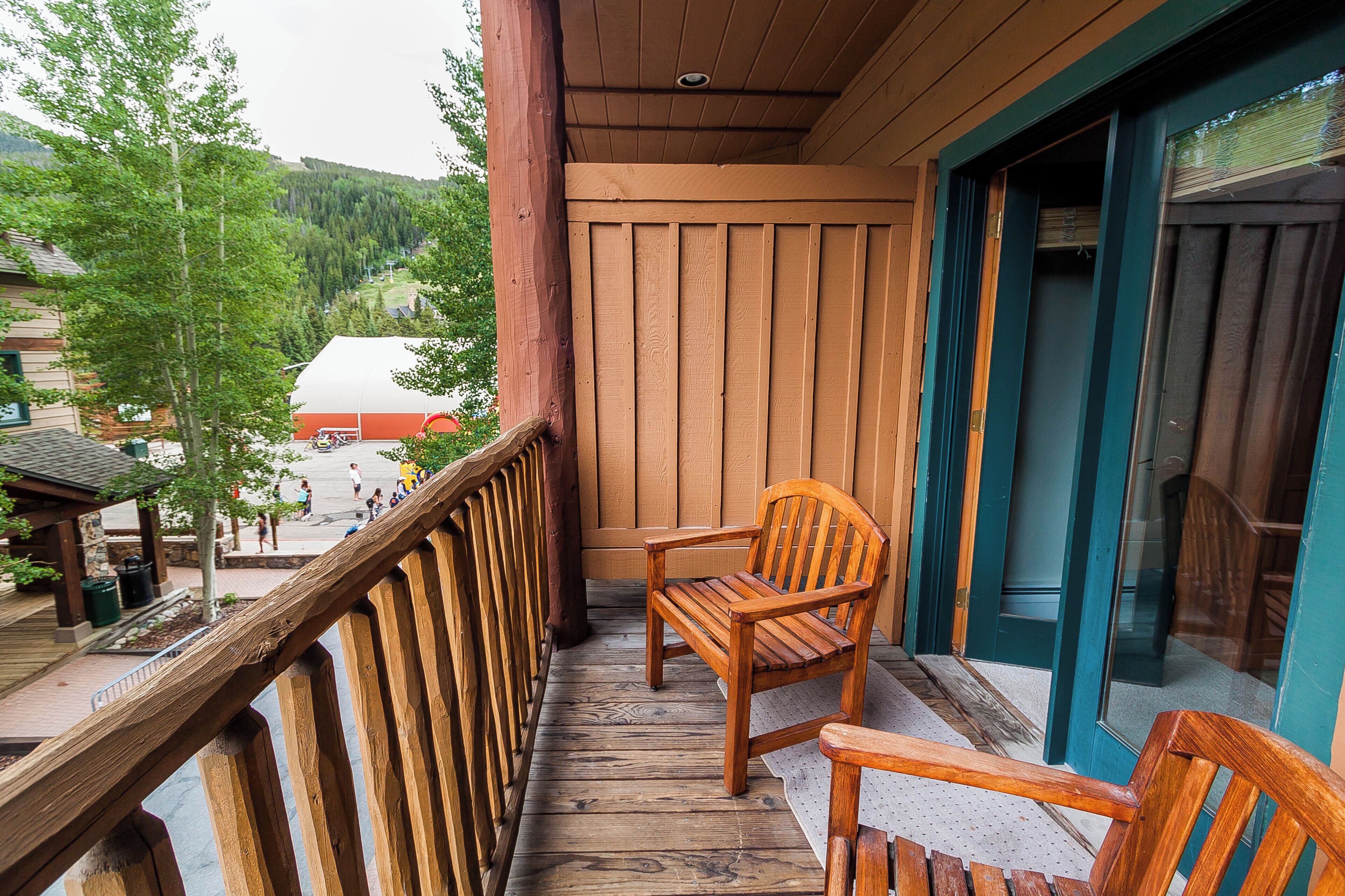 A wooden deck with a wooden chair and a bench, overlooking a building with a red roof and a view of trees and hills.