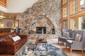 A stone fireplace with a wooden mantel, a leather sofa, a wooden coffee table, and two armchairs in a living room with large windows. - Expedition Station #8599 (Keystone)