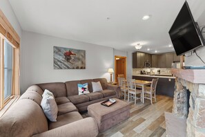 A living room with a sofa, ottoman, and a painting on the wall. There is a dining area with a table and chairs, and a kitchen area with a counter and appliances. - Expedition Station #8599 (Keystone)