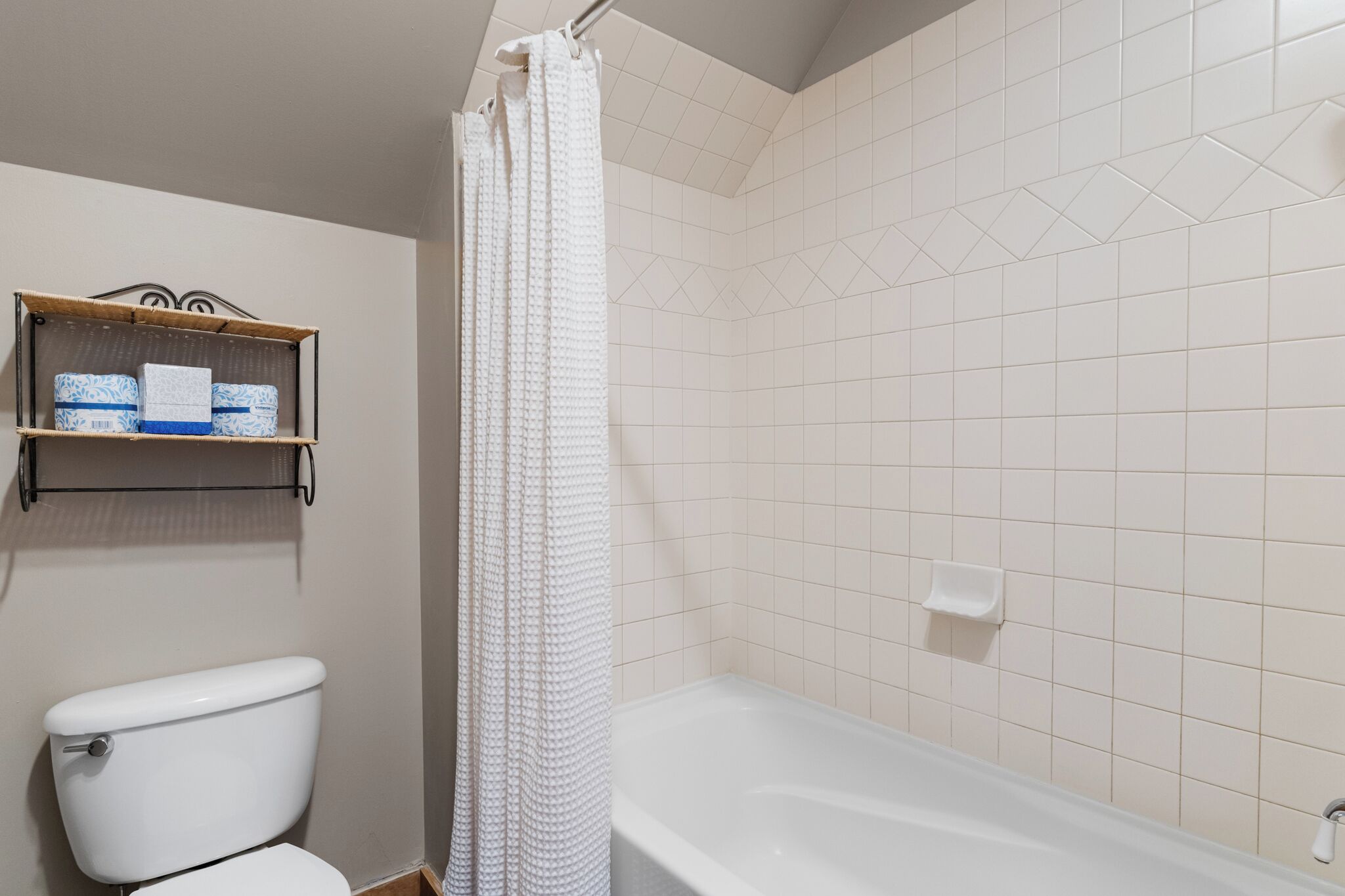 A bathroom with a toilet, a bathtub, and a shower curtain. There is a shelf with toiletries above the toilet.